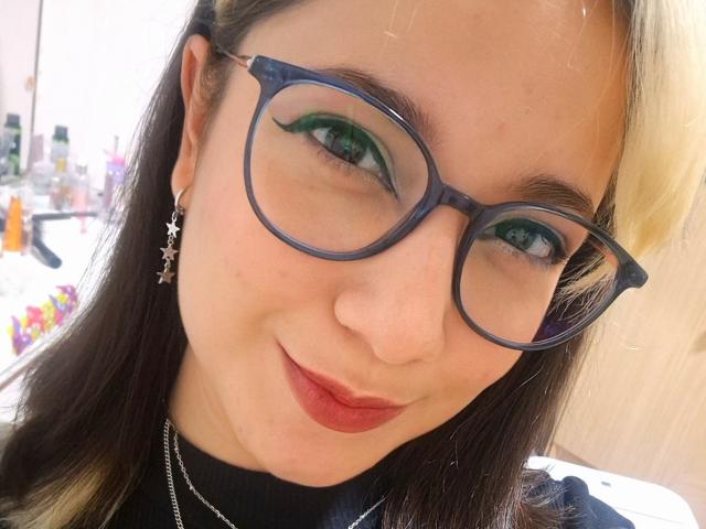 ErinFoxy has green eyeshadow, black-rimmed glasses, and red lipstick, smiling with blonde and brown hair, wearing star earrings and a black turtleneck.