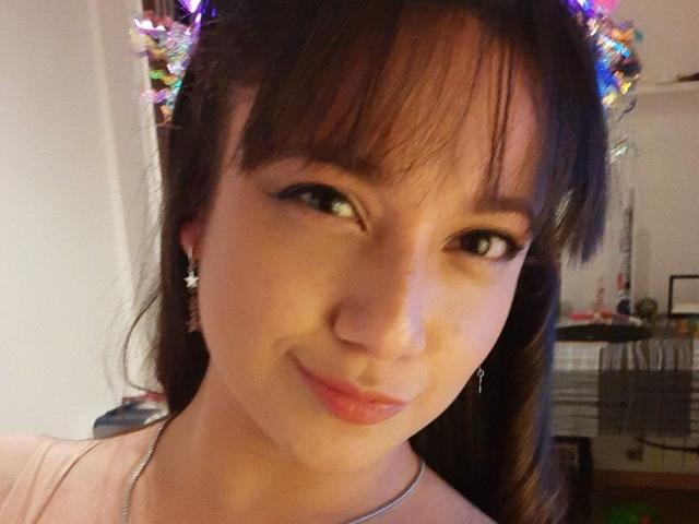 ErinFoxy with dark brown hair, bangs, and a subtle smile, wearing star earrings, and a pink top, winking with a cute, slightly mischievous look, and a silver necklace.