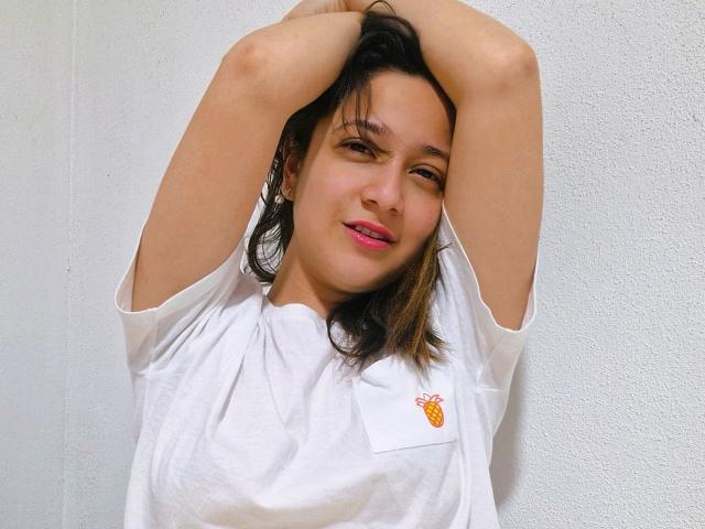 ErinFoxy with arms above head, wearing white shirt, pink lipstick, brown hair, and small orange logo on shirt, smiling slightly, standing against white wall.