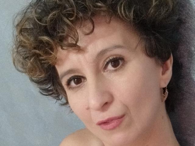 NikHotty has curly brown hair, light skin, brown eyes, and pink lipstick. She's wearing small earrings and has a subtle smile. Her boobs are not visible.