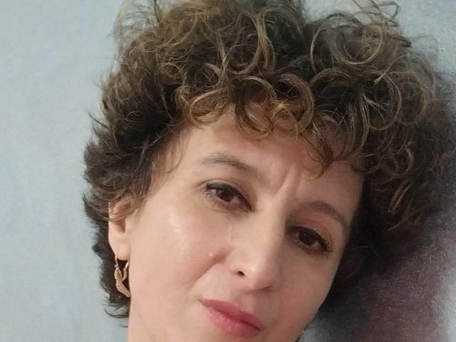 NikHotty has curly brown hair, light skin, brown eyes, and pink lips, wearing small gold earrings, with a slight smile, looking directly at the camera.