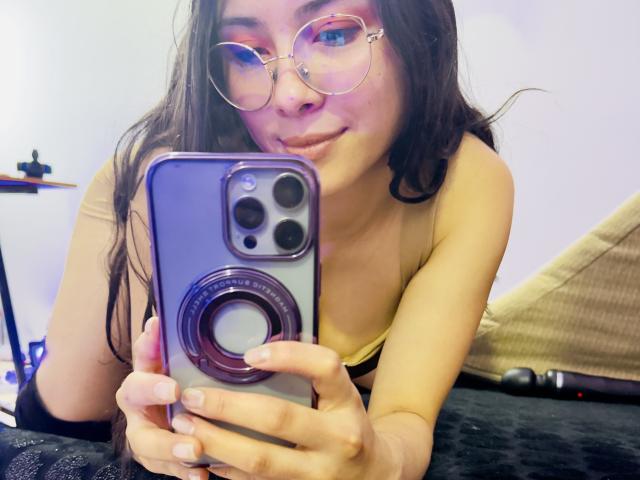 Isabella, una principessa con gli occhiali, ombretto rosa e lunghi capelli castani, sta scattando un selfie con un telefono cellulare viola, mostrando il suo seno piccolo, indossando una maglietta nera..