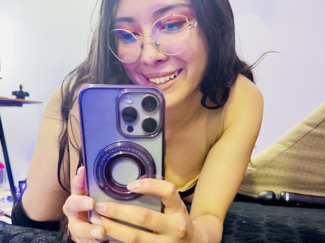 IsabellaPrincess with long brown hair, glasses, smiling, holding a phone with a pop-up camera, wearing a yellow top, showing her tits, taking a selfie.