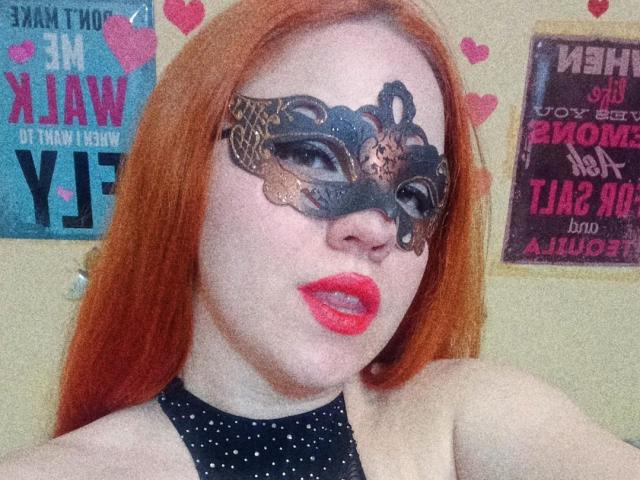 SpicyGrace with red hair, wearing a black lace masquerade mask, red lipstick, and a black polka dot halter top, showing off her pale skin and sexy face.