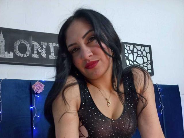 LanaBox with long black hair, red lipstick, and a sheer black dress with sparkles, showing her small tits, wearing a gold necklace, sitting against a blue wall.