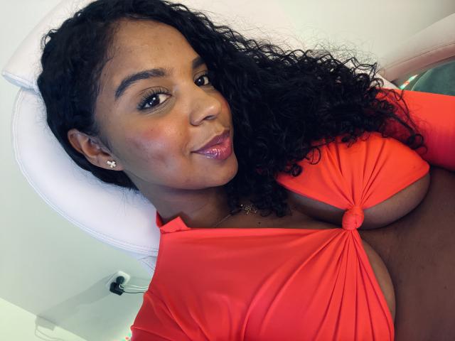Lisa Mendez mit dunklen, lockigen Haaren, trägt ein rotes Oberteil mit Schluppe, das ihre großen Brüste zeigt, lächelt mit pinkem Lippenstift, im Hintergrund ein weißer Stuhl, weiches Licht auf ihrer dunklen Haut..