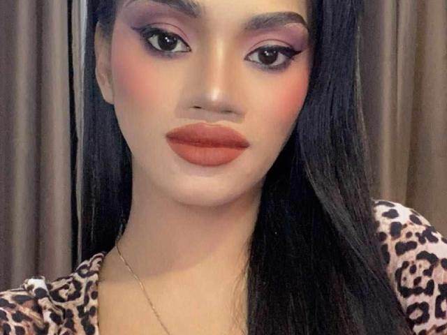 SevenInchesCockTS tem um rosto atraente, com olhos escuros, sombra rosa, sobrancelhas grossas e batom laranja. Ela está vestindo uma blusa com estampa de leopardo e tem cabelos longos e pretos..