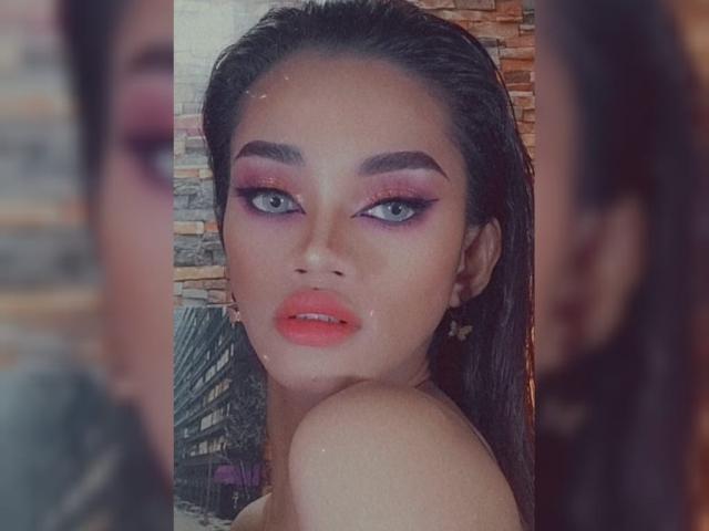 A SevenInchesCockTS tem cabelos escuros e úmidos, sombra rosa, delineador preto marcante, batom vermelho e uma expressão séria em uma selfie no espelho. Seus seios estão ligeiramente visíveis..