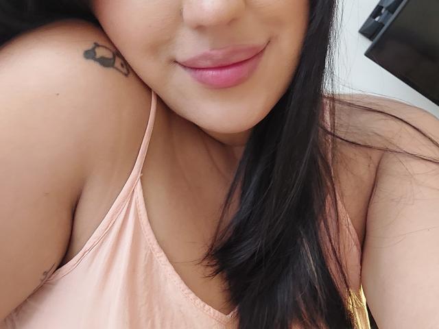 LauraRichis with pink lips, black hair, and a tattoo on her shoulder, wearing a pink tank top, showing her bare shoulder and upper chest. Boobs visible, smiling slightly.