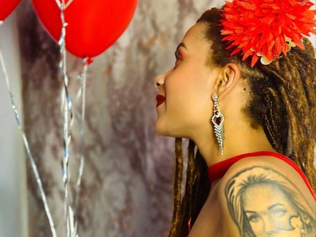 KendallQuinn with red lipstick, red earrings, red flower in hair, red top, and detailed tattoo on right shoulder, looking at red balloons. Her nose is pierced.