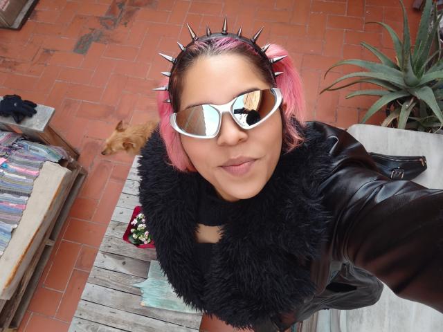 Chili está recebendo um footjob, vestindo óculos de sol reflexivos brancos, faixa de cabelo com pontas, jaqueta de couro preta e lenço peludo, com gato nas proximidades.
