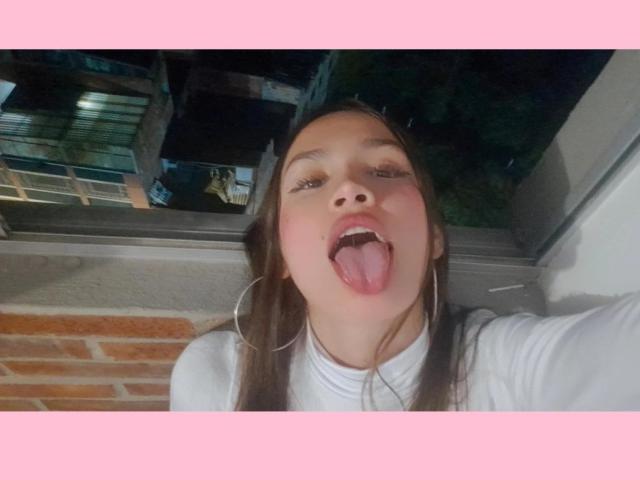 LiliMoore has a playful expression, tongue out, with brown hair, hoop earrings, and a white turtleneck. Her face is centered, with a pink border on the image.