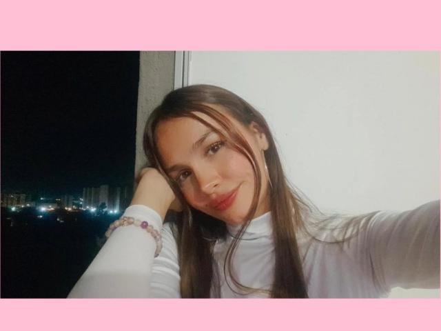LiliiMoore with long brown hair, wearing a white turtleneck, resting her head on her hand, smiling slightly, pink border, city lights in the background, bracelet on her wrist.