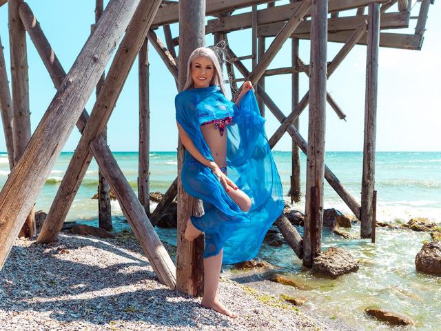 KassandraGray stands under a wooden pier, holding a blue sheer scarf that reveals her small, perky tits and bare ass. She's smiling, with a colorful bikini top visible.