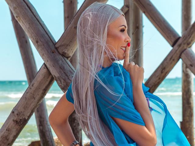 KassandraGray with long silver hair, blue sheer top, red lipstick, and a finger to her mouth, standing by wooden cross-beams, ocean in the background.