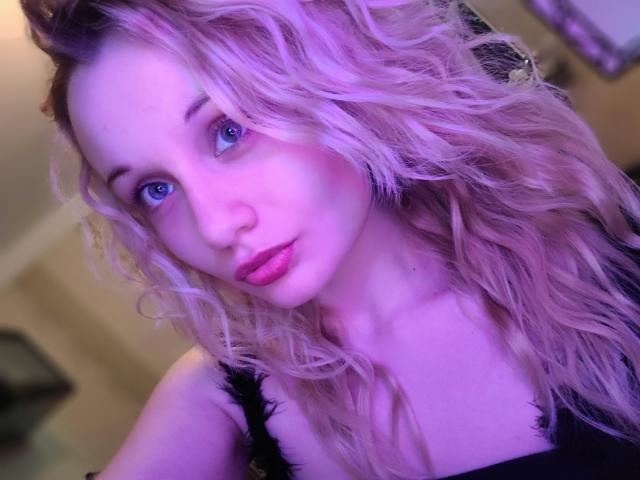 CherylGalaxy with pink wavy hair, blue eyes, pink lipstick, and black lace top, looking at camera, purple lighting, soft skin, slightly parted pink lips.