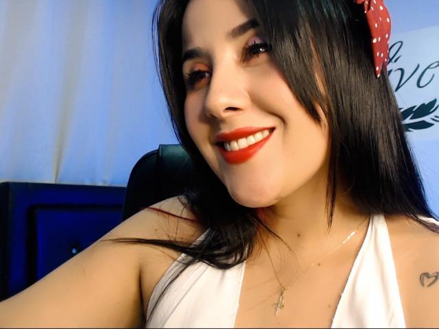 CameronGrey, con capelli scuri, rossetto rosso, top bianco con spalline, molletta per capelli a pois rossi, sorridente, che mostra il seno, piccolo tatuaggio sulla spalla destra, collana, seduta di fronte a uno schermo blu..