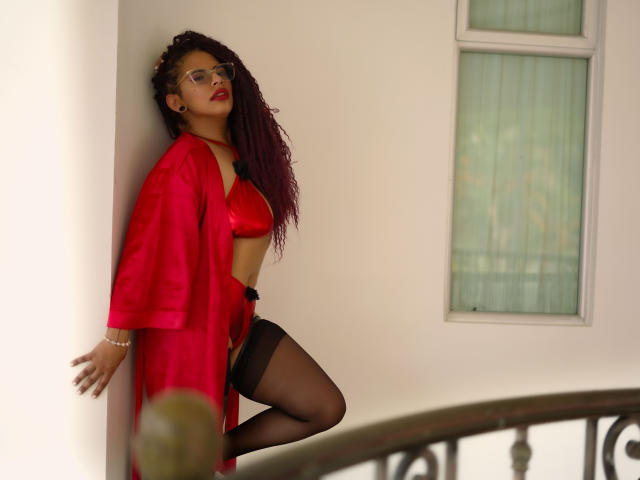 Karyn Black mit lockigem, rotem Haar, einem roten Seidenbademantel, einem schwarzen BH und Unterhose, schwarzen Strümpfen und einer Brille, lehnt an einer Wand, zeigt ihre Brüste und ihre Vagina, steht auf einem Balkon..