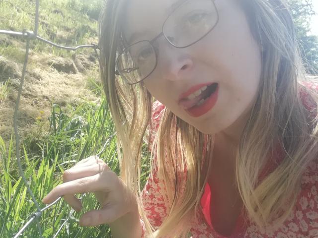  AlexandraSweet with glasses, blonde hair, red lips, open mouth, pink floral shirt, holding grass, sunlight, slightly messy hair, looking down, right hand near her face.