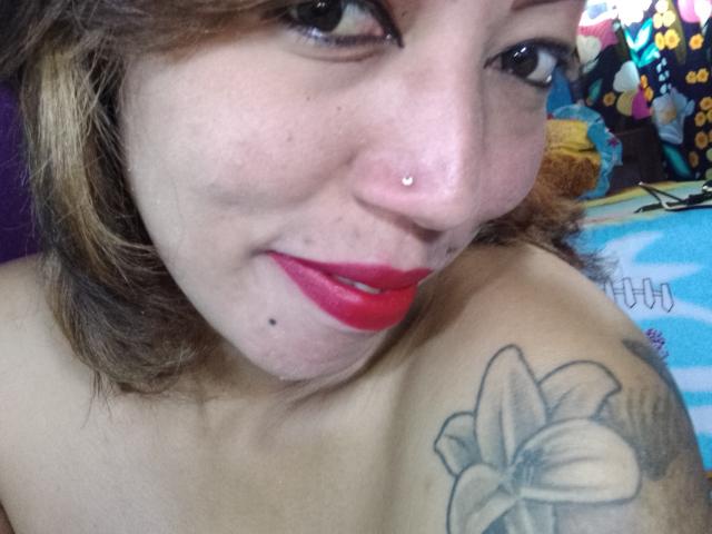 AnniaTattoo with red lipstick, nose ring, and tattooed shoulder showing a large flower, smiling at the camera, topless, brown hair, freckles, and a small mole under her right lip.