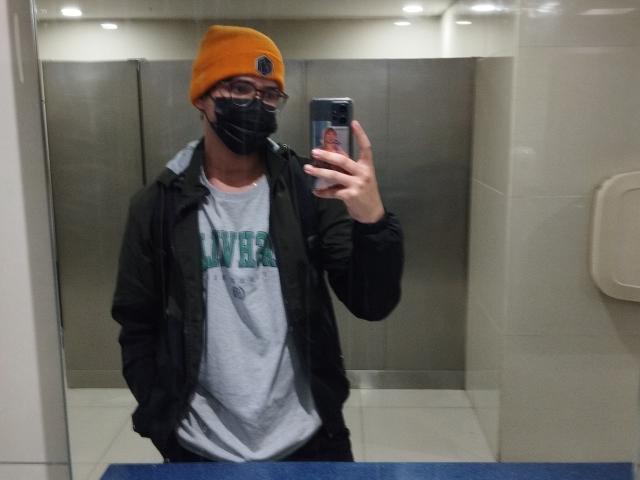 EddTerence en un selfie frente a un espejo de baño, usando un gorro amarillo, una máscara negra, gafas, una camiseta verde y gris con el logo de "UNICEF", una chaqueta negra, sosteniendo un teléfono, con las manos en los bolsillos..
