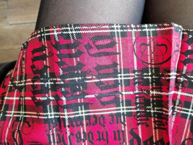 De rode geruite rok van AlexandraSweet, met zwarte en gele lijnen en een patroon dat lijkt op graffiti, is gedrapeerd over haar zwarte panty's. De textuur van de rok is licht ruw..