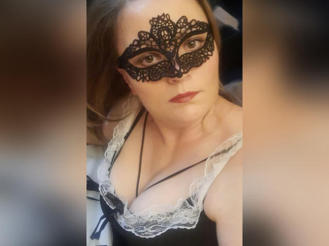 ArtMasterX, com uma máscara de renda preta, seios médios e uma blusa preta com detalhes em renda, tem lábios vermelhos e cabelo castanho, olhando para a câmera com uma expressão séria..
