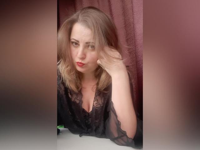 ArtMasterX, com cabelo loiro, batom vermelho e blusa preta transparente, fazendo um leve biquinho, tocando a cabeça com uma mão, em frente a uma cortina rosa desfocada..