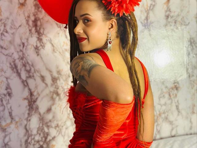 Kendall Quinn con lunghi dreadlocks, vestita di rosso, con guanti rossi, orecchini rossi e un fiore rosso tra i capelli, sorridente, che mostra un tatuaggio sulla spalla destra, con rossetto rosso, e che guarda oltre la spalla..