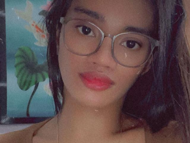 SevenInchesCockTS con gafas, cabello oscuro y lápiz labial rojo, mirando directamente a la cámara. Tiene una cara linda, muy linda, y una nariz pequeña y bonita. Sus pechos son pequeños..