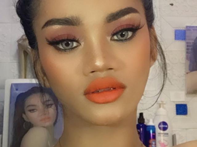 SevenInchesCockTS tiene lápiz labial naranja, delineador de ojos negro grueso y cejas arqueadas. Está mirando a la cámara con ojos grises, y su cabello oscuro está recogido..