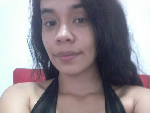 ViiviBush has medium brown skin, black wavy hair, small nose, and is smiling slightly. She's wearing a black halter top, showing her bare shoulders and small tits.