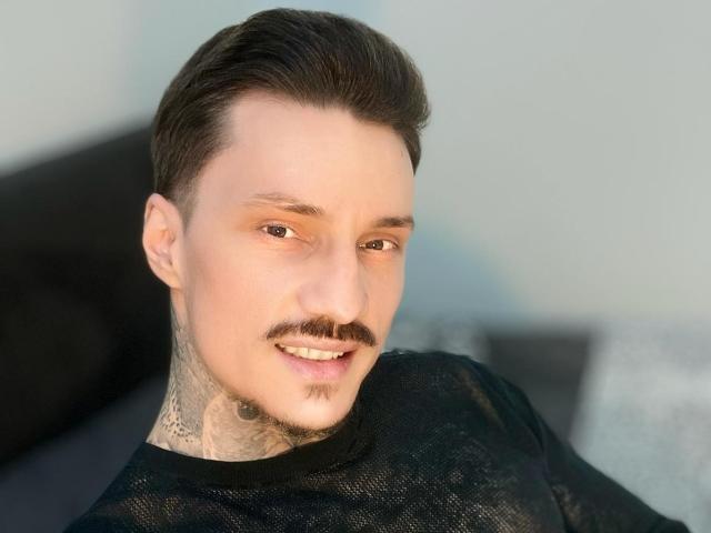 TyagoDoll has a mustache, tattoos on his neck, and is smiling with short dark hair, wearing a black mesh shirt. His skin is fair, and he's looking to the side.