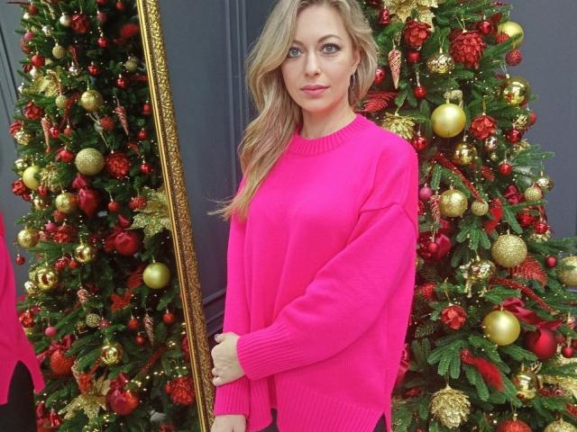 SweetLadyXX with blonde hair, pink sweater, standing in front of two decorated Christmas trees, holding a gold-framed mirror, looking at camera, soft smile, cute, festive, attractive.