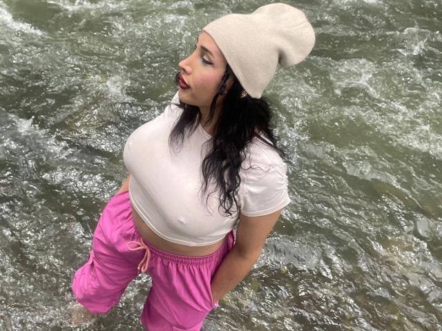 PamelaBigCoCk with wet white tee, pink pants, and beige beanie, standing in flowing water, showing her big tits and black hair, looking to the side with red lipstick.
