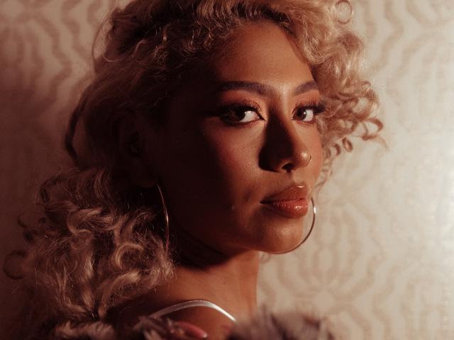 LilithWalker's face with curly blonde hair, dark skin, hoop earrings, and glossy pink lips, looking to the side with a subtle, confident smile. Warm, soft lighting highlights her features.