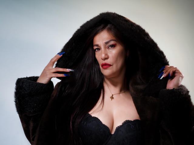 MagikGodess avec de longs cheveux noirs, du rouge à lèvres rouge et des ongles bleus, portant un soutien-gorge en dentelle noire et un manteau marron duveteux, tenant la capuche du manteau. Ses seins sont visibles..