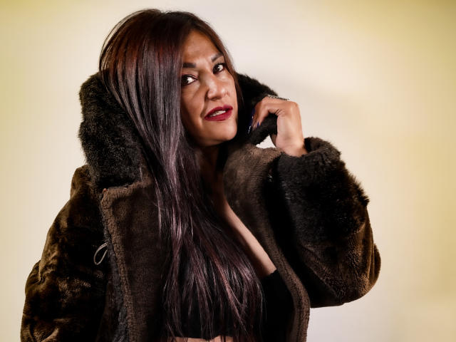 MagikGodess with long dark hair, red lipstick, and brown fur coat, touching her face, smiling, showing off her tits slightly through the coat, against a plain yellow background.