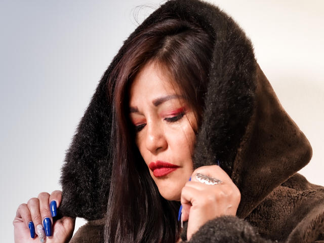 MagikGodess mit dunkelbrauner Fellhaube, rotem Lippenstift, blauen Nägeln und einem silbernen Ring, die Haube hochgehalten, Augen geschlossen, weiches Licht, leichtes Stirnrunzeln, Halbtotale, realistisch..