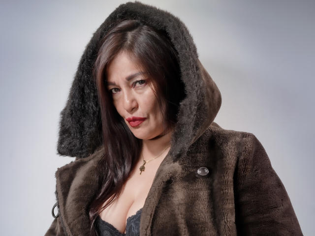 MagikGodess with dark brown hair, red lipstick, wearing a brown fur hooded coat, showing cleavage in a gray lace top, looking slightly annoyed.