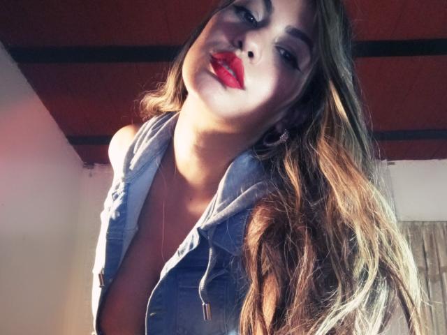 AnastaciaMonaco with long brown hair, red lipstick, and blue denim shirt, showing a bit of her tits, looking down with a slight smile, sunlight on her face.