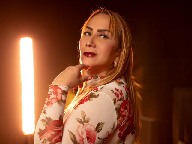 DianeOasis, blonde woman, floral dress, red lipstick, hand on chin, looking over shoulder, warm light, medium shot, soft smile, visible tits, earrings, dark background, illuminated vertical light.