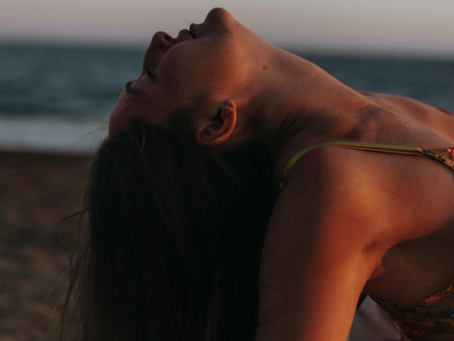 BiancaHotLove with her head tilted back, long brown hair falling, wearing a yellow-strapped top, showing her tits, and a sun-kissed, muscular neck at sunset.