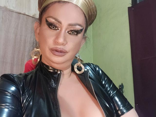 MistressKinykyJulia with shiny black latex top, gold headband, heavy eyeliner, and choker, showing cleavage, wearing large hoop earrings, and sporting a serious expression.