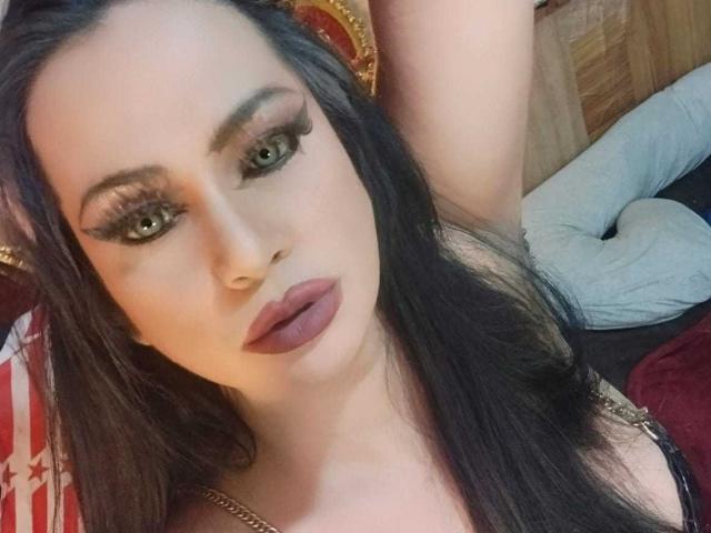 MistressKinykyJulia with long black hair, green eyes, and thick eyelashes, looking at the camera with full, purple-lipped, slightly parted mouth, and her arm raised. Boobs visible.