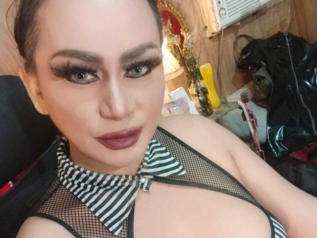 MistressKinykyJulia with dark lipstick, heavy eyeliner, and long lashes, wearing a black and white striped, mesh top, exposing her large, round tits. She has a confident, sexy look.