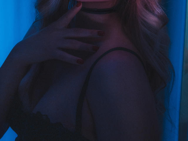 BiancaHotLove with her hand on her tits, wearing a black choker and lace bra, blue lighting, wavy blonde hair, red nails, and a sexy, dark, moody vibe.