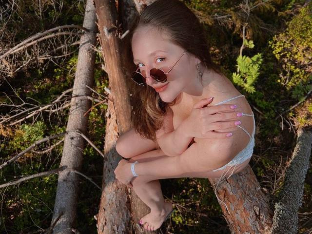 BiancaHotLove, with small tits, sitting on a tree, wearing round sunglasses, white lace bra, and anklet, in a forest, smiling, hands on chest, pink nails, brown hair.
