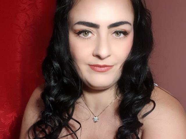 MistressMayaDiamondXx hat helle Haut, grüne Augen, schwarzes, welliges Haar und lächelt mit rotem Lippenstift. Sie trägt eine kleine silberne Halskette mit einem Totenkopf-Anhänger..