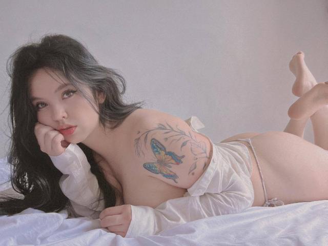 ElyMoon with black hair, lying on white bed, wearing an open shirt, revealing her tits and a colorful tattoo of blue butterflies on her right breast, legs up.